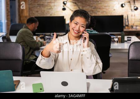 Asian freelancer gesturing thumbs up while talking on phone in coworking space Stock Photo