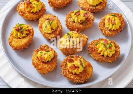 close-up of Deep fried deviled eggs with crispy coating of panko and parmesan, sprinkled with fresh chives and crumbled bacon on white plate on wooden Stock Photo