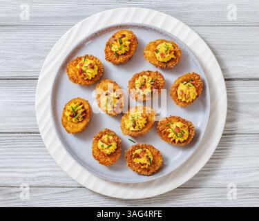 Deep fried deviled eggs with crispy coating of panko and parmesan, sprinkled with fresh chives and crumbled bacon on white plate on wooden table with Stock Photo