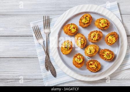 Deep fried deviled eggs with crispy coating of panko and parmesan, sprinkled with fresh chives and crumbled bacon on white plate on wooden table with Stock Photo