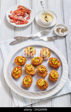 Deep fried deviled eggs with crispy coating of panko and parmesan, sprinkled with fresh chives and crumbled bacon on white plate on wooden table with Stock Photo