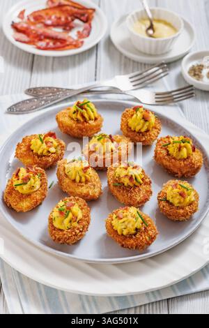 Deep fried deviled eggs with crispy coating of panko and parmesan, sprinkled with fresh chives and crumbled bacon on white plate on wooden table with Stock Photo