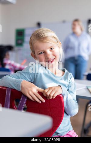 Smiling girl sitting on chair in school classroom, looking at camera Stock Photo