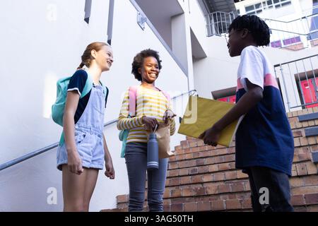 Talking and smiling, three diverse students with backpacks standing on school stairs Stock Photo
