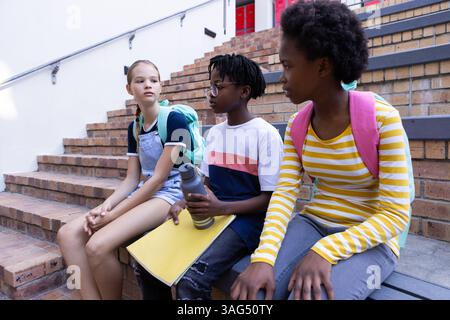 Sitting on stairs, three diverse students with backpacks talking after school Stock Photo