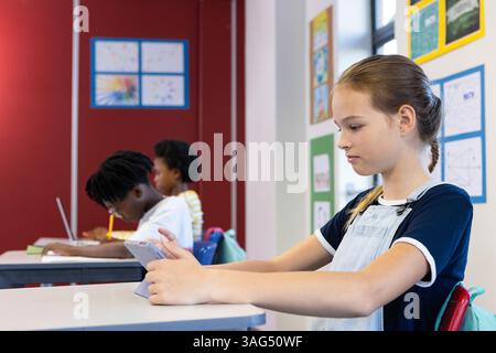 In school, girl using tablet while classmates studying at desks in classroom Stock Photo