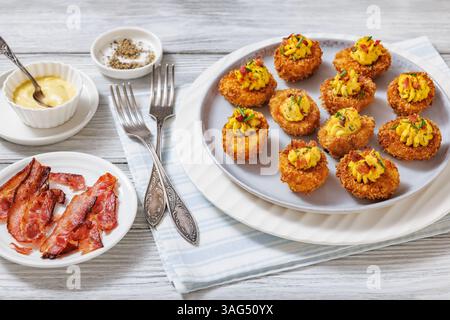 Deep fried deviled eggs with crispy coating of panko and parmesan, sprinkled with fresh chives and crumbled bacon on white plate on wooden table with Stock Photo