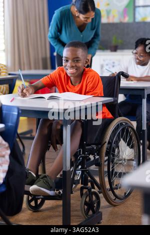 In school, boy in wheelchair writing in notebook, smiling in classroom Stock Photo
