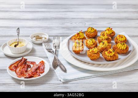 Deep fried deviled eggs with crispy coating of panko and parmesan, sprinkled with fresh chives and crumbled bacon on white plate on wooden table with Stock Photo