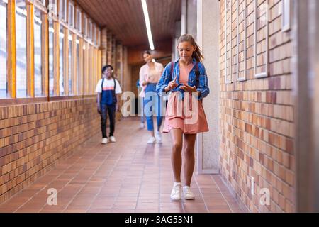 Walking in school hallway, girl using smartphone, students in background, copy space Stock Photo