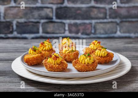 Deep fried deviled eggs with crispy coating of panko and parmesan, sprinkled with fresh chives and crumbled bacon on plate on table with brick wall in Stock Photo