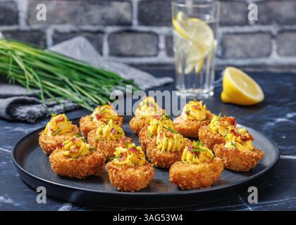 Deep fried deviled eggs with crispy coating of panko and parmesan, sprinkled with fresh chives and crumbled bacon on dark grey plate on table with bri Stock Photo