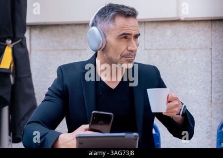 Mature man with headphones using devices and drinking coffee at outdoor cafe Stock Photo