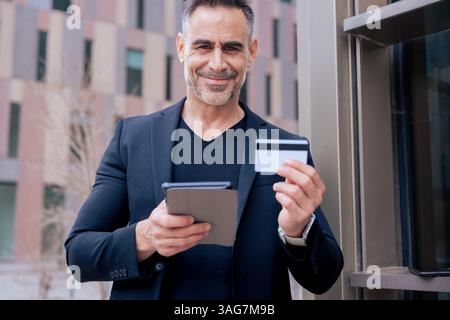 Mature man smiling while holding credit card and tablet outdoors in city Stock Photo