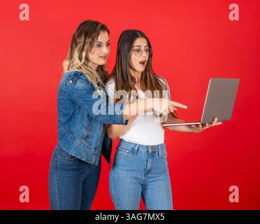 Two women using laptop together, pointing at computer screen over red studio background. Shocked face expression. Attractive amazed trendy girls. Stock Photo