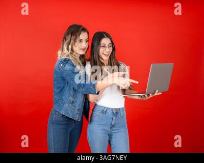 pointing at computer screen over red studio background. Shocked face expression. Attractive amazed trendy girls wear fashionable denim jacket and jean Stock Photo