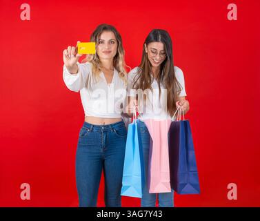 two beautiful caucasian women standing over red background. Blonde girl showing credit card smiling to camera. Holding and looking into shopping bags Stock Photo