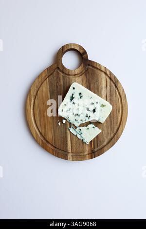 Round rustic wooden cheese board with a piece of blue cheese, cracked and crumble. Top view, neutral background Stock Photo