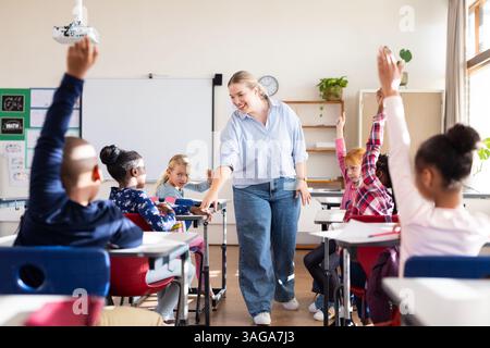 In school, female teacher engaging with diverse students raising hands ...