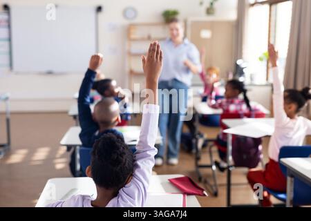 In school, diverse students raising hands to answer female teacher's question in classroom Stock Photo