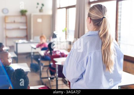 In school, female teacher standing in classroom, diverse students sitting at desks listening Stock Photo