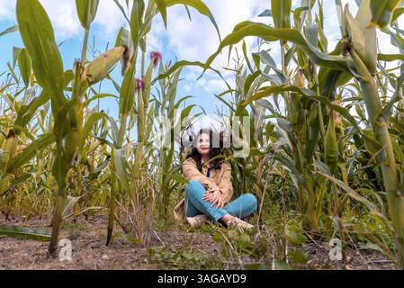 Funny concept with a young woman sitting in the middle of a corn field with her curly hair hang on the corn leaves Stock Photo