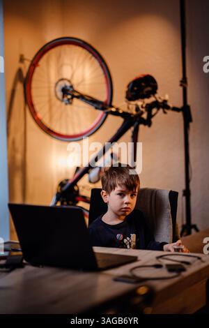 Focused boy sitting at a desk with a laptop in a modern room, bicycle mounted on wall in background, warm lighting Stock Photo