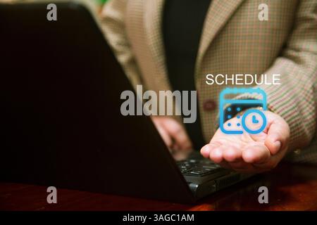 A business person in a checkered blazer works on a laptop, while holding out their hand with a digital calendar and clock icon floating above it, symb Stock Photo