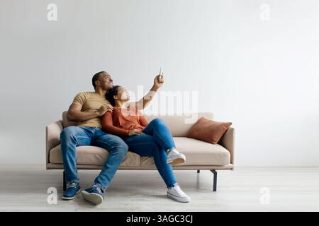 Split system. Cheerful young black couple with remote relaxing under air conditioner Stock Photo