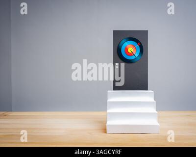 Big modern colorful target dartboard sign at the door at top of white steps, isolated on wooden table, grey background with copy space. Business goal, Stock Photo