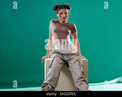 Young woman in casual outfit posing confidently on cube in studio. Fashion, style, confidence, female, model, contemporary Stock Photo