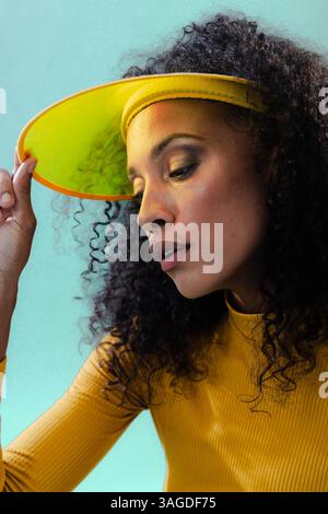 Woman wearing yellow visor and sweater posing confidently against teal background. Fashion, style, confidence, portrait, trendy, colorful Stock Photo