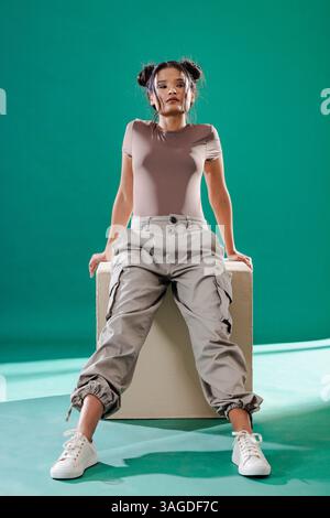 Young woman in casual outfit sitting confidently on box in studio. Fashion, confidence, model, posing, modern, lifestyle Stock Photo