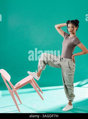 Confident woman in casual outfit balancing on pink chair in studio. fashion, confidence, lifestyle, creative, posing, contemporary Stock Photo