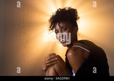Smiling woman with curly hair sitting wrapped in cozy black sweater. Portrait, comfort, warmth, fashion, joyful, curly hairstyle Stock Photo