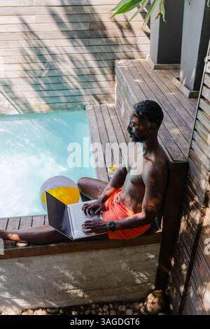 Man in red shorts using laptop by pool, enjoying sunny morning outdoors. Technology, relaxation, leisure, summer, workation, wireless Stock Photo
