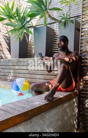 Relaxing by pool, African American man in orange shorts using smartphone, enjoying leisure time. relaxation, vacation, technology, summer, swimming, m Stock Photo