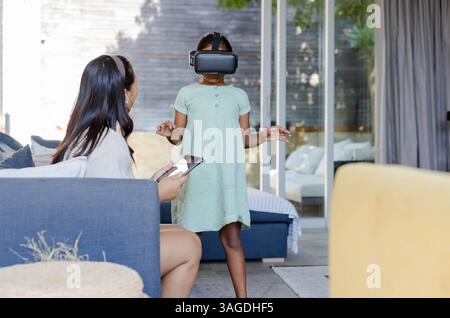 African American girl using VR headset while mother watches with smartphone at home. Family, virtual reality, bonding, technology, entertainment, digi Stock Photo