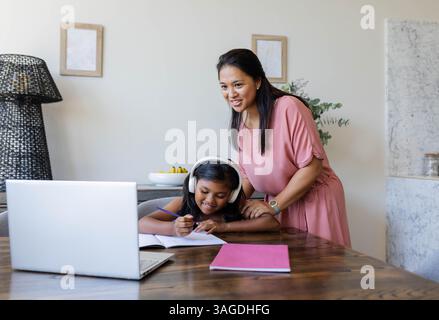 Mother helping daughter with homework at home, smiling and using laptop. education, tutoring, bonding, family, technology, schoolwork Stock Photo