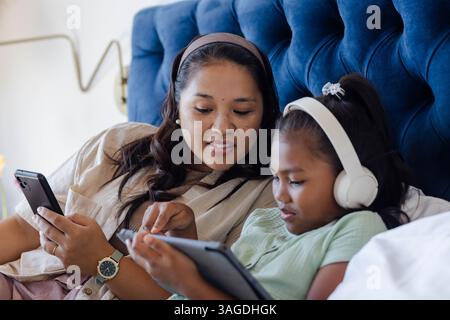 Mother and daughter relaxing on bed, using tablet and smartphone together. Family, bonding, technology, relaxation, leisure, parenting Stock Photo
