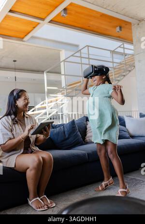 Asian mother using tablet while daughter enjoys VR headset in living room. Family, technology, bonding, interaction, leisure, digital Stock Photo