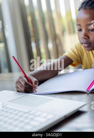 African American boy writing in notebook at home, focusing on homework near laptop. studying, learning, education, homeschooling, concentration, techn Stock Photo