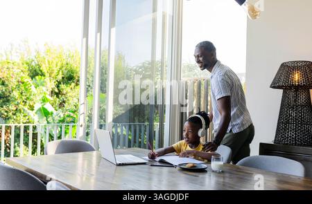 African American father helping daughter with homework at home, using laptop and headphones. Parenting, tutoring, technology, education, family, guida Stock Photo