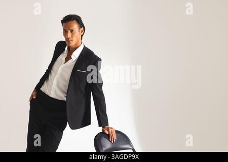Confident man in black suit leaning on chair, posing in fashion studio. style, elegance, confidence, photoshoot, formal, attire Stock Photo