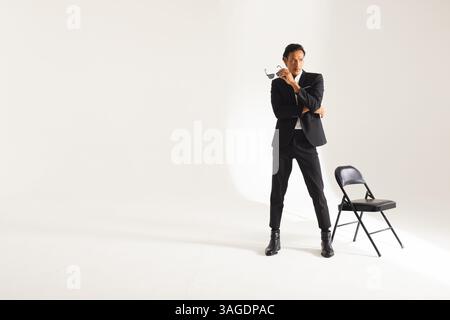 Confident man in stylish black suit posing with sunglasses near chair. Fashion, confidence, style, elegance, classic, dapper Stock Photo