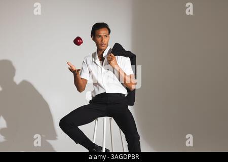 Stylish man in white shirt tossing apple while sitting on stool in studio. Fashion, minimalism, casual, trendy, elegance, posing Stock Photo