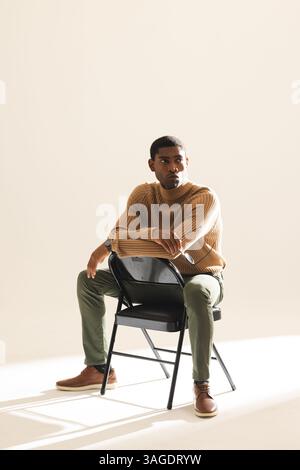 African American man in stylish outfit sitting on chair, looking thoughtfully ahead. Fashion, style, contemplation, modern, trendy, elegance Stock Photo