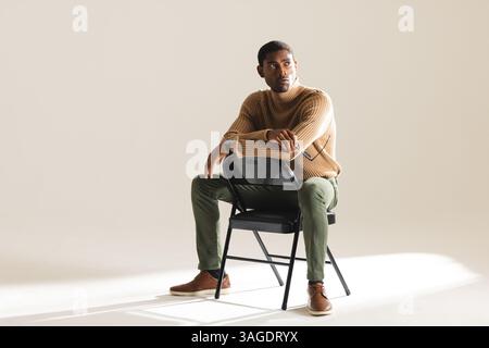 Stylish man in sweater sitting on chair, looking thoughtfully in studio. Fashion, contemplation, portrait, male, style, winter Stock Photo