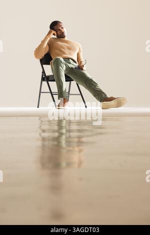 Sitting on chair, African American man in stylish outfit holding glasses thoughtfully. Fashion, style, contemplation, eyewear, thinking, urban Stock Photo