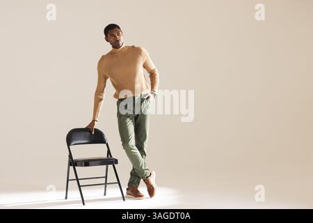 Confident man in stylish outfit leaning on chair in modern studio. Fashion, confidence, style, elegance, photography, portrait Stock Photo